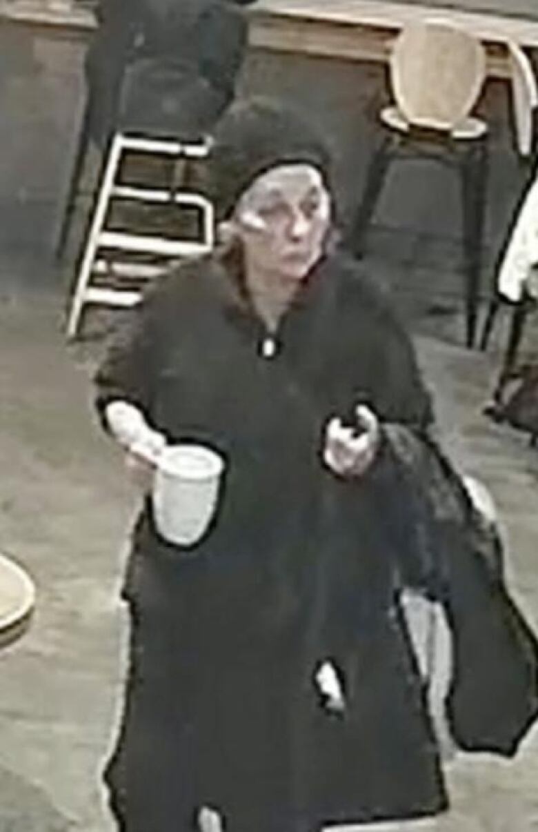 A woman in a black beanie and black sweater holds a coffee cup in one hand and a black jacket in the other. 