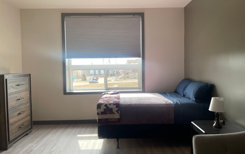 A photo shows the end of a suite, with a dresser on the left side and a bed on the right. The blinds on the window are partially pulled up, letting sunlight in. 