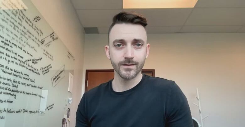 A man in a black t-shirt speaks on Zoom in a room with a white board. 