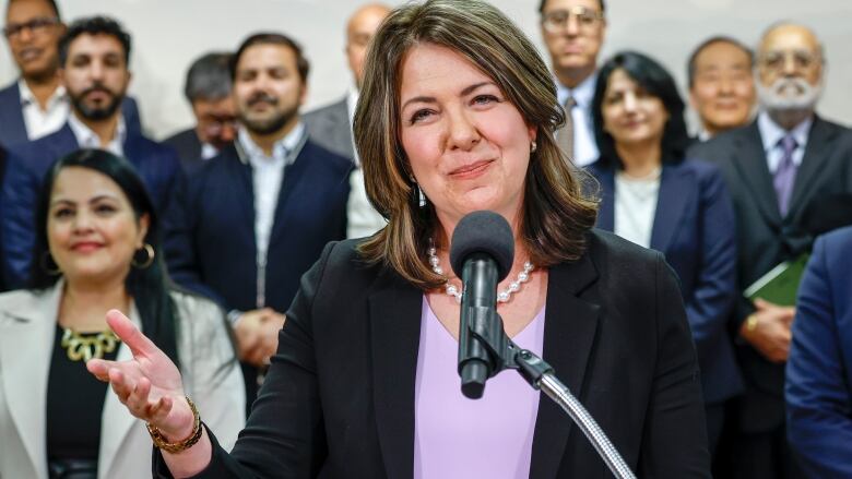 A woman stands behind a microphone and smiles. A crowd of people stands behind her.