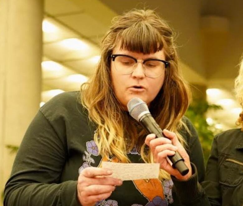 Juls Budau speaks in to a microphone held by another person, wearing glasses and a cue card to read from. 