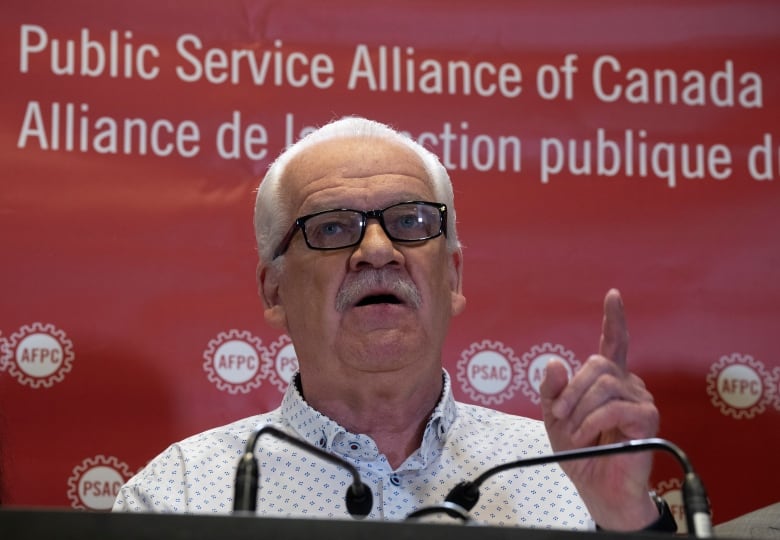 A man raises a finger while speaking at a lectern.