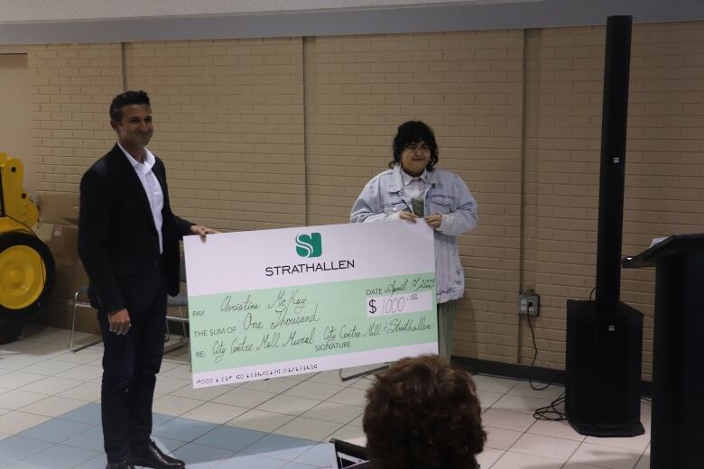 A man hands a woman a green cheque that reads 'Strathallen' for $1,000