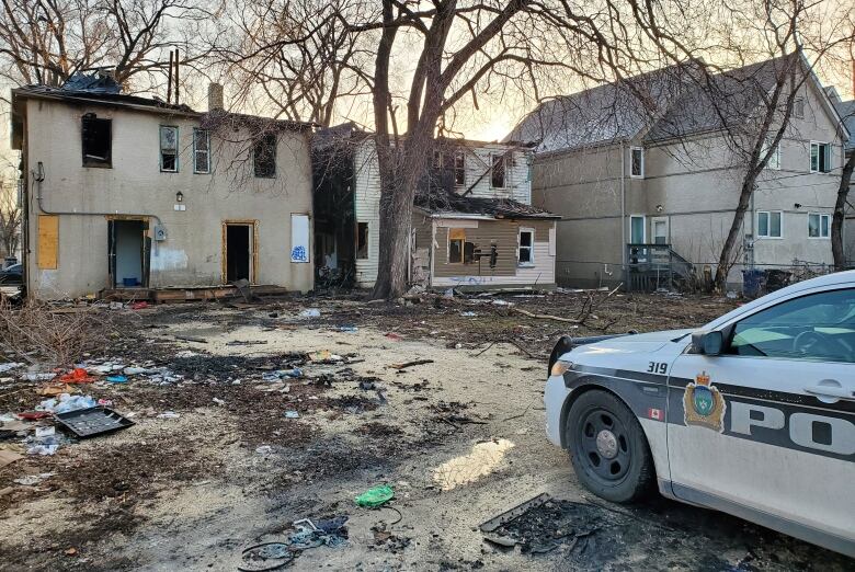 A police car in the right foreground is parked in front of two two-storey residences that were burned in a fire. The windows are broken and doors open on both buildings.