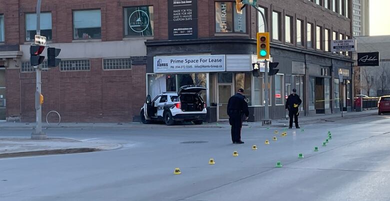 Police officers set down markers in an intersection at the scene of a crash. A police cruiser is in the background, its doors open, with the front end smashed into a building.