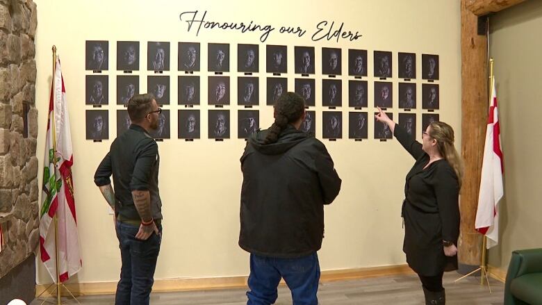 Three people look at a wall filled with black and white portraits of elders from the community