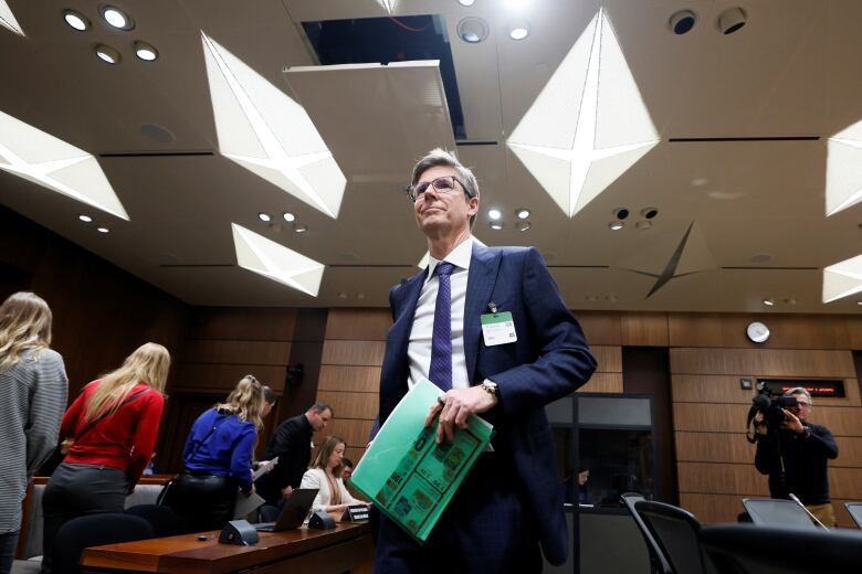 A man in a suit stands in a conference room, holding some papers. 