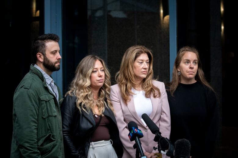 Four people with solemn expressions stand together outside a glass window on an overcast day.