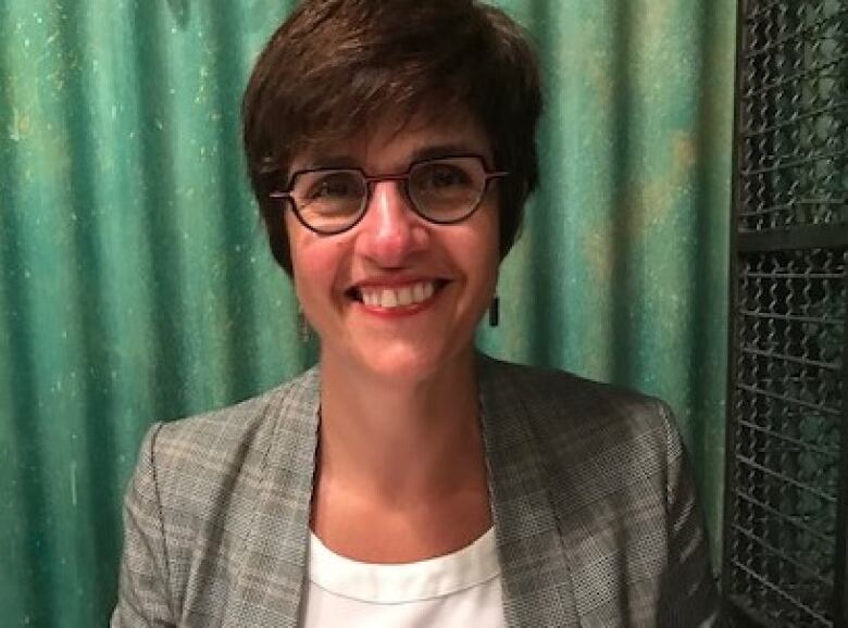 A woman with short hair and glasses smiles in front of a green background.