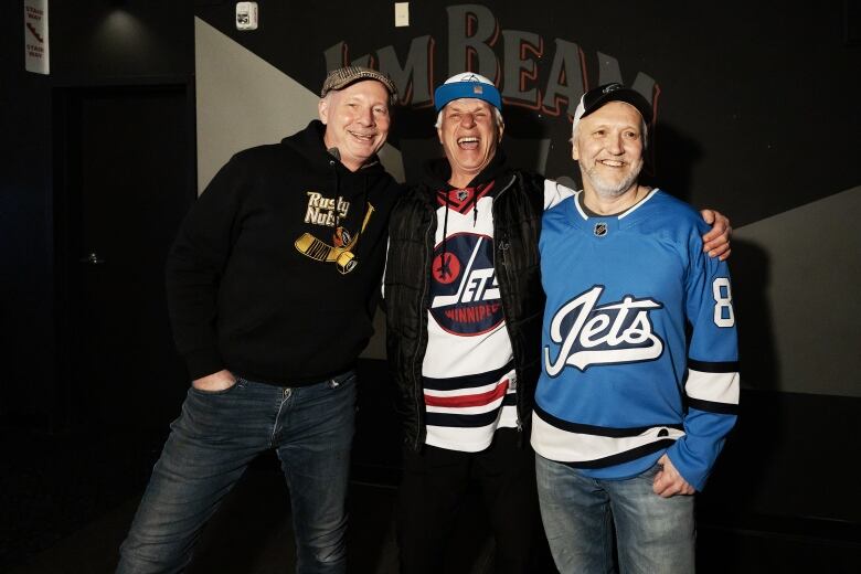 Three middle-aged men in hockey jerseys smile as they stand arm-in-arm.