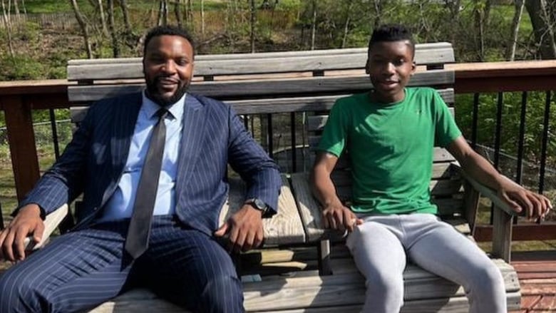 A Black man in a suit and a Black teen sit together on a bench smiling. 