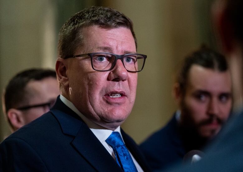 A man with glass and a blue tie speaks to someone across the photo frame.