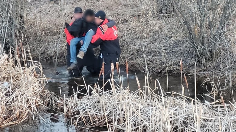 Men in drysuits carry a man in jeand and a hoodie across a stream.