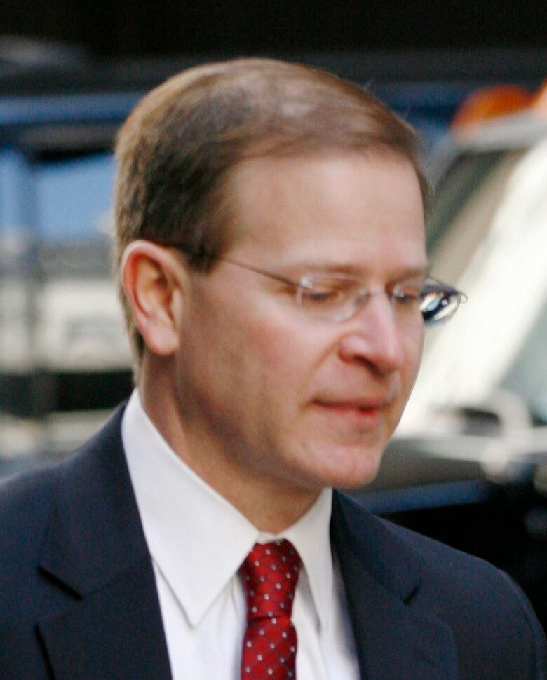 A man with short brown hair and glasses wearing a dark suit and red tie carries a stack of papers under his arm. 