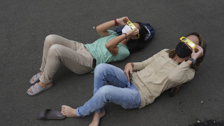 Two people are shown lying on pavement wearing special glasses and looking skyward.