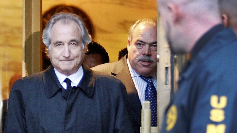 Bernard Madoff leaves U.S. District Court wearing a navy trench coat, white collared-shirt and black tie. He is walking through a door that is open for him with two US Marshall guards standing outside to his left.