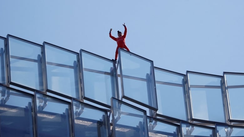 A man in red stands atop a glass building holding his hands above his head in triumph. 