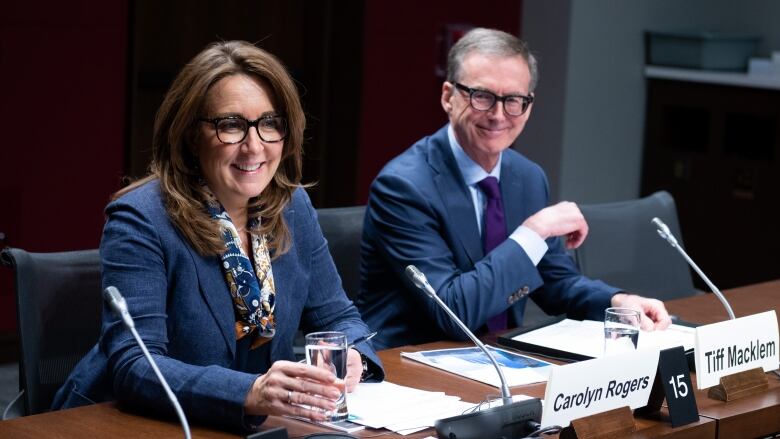 A man and a woman wearing blue suits and smiling sit at a table before microphones.