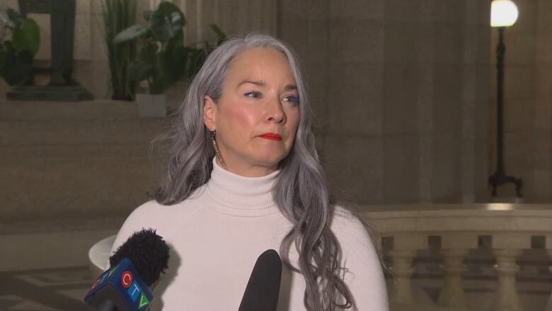 A woman wearing a white sweater speaks to media while standing behind two cameras.