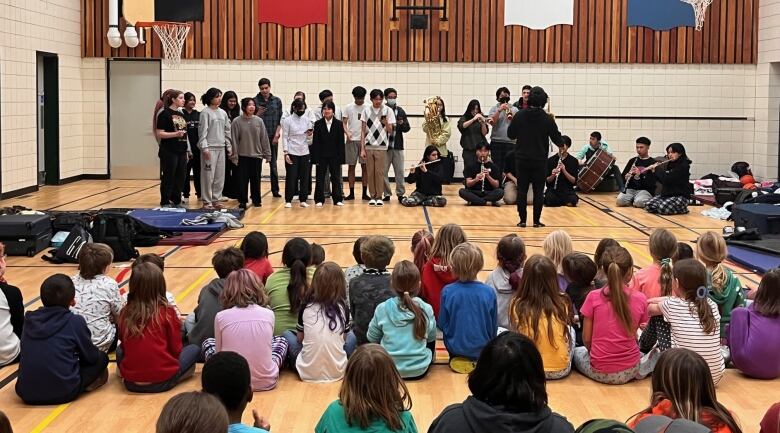 A group of high school students are shown singing and playing musical instruments for a group of elementary school students.