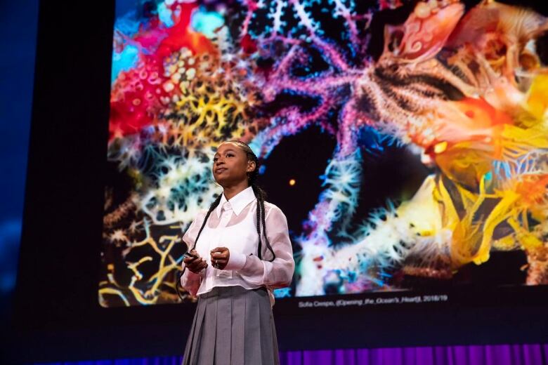 A woman stands on a stage in front of a colourful collage image displayed on a screen.