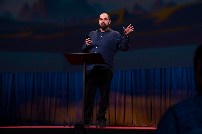 A man on a stage speaks to a crowd with his hand outstretched.