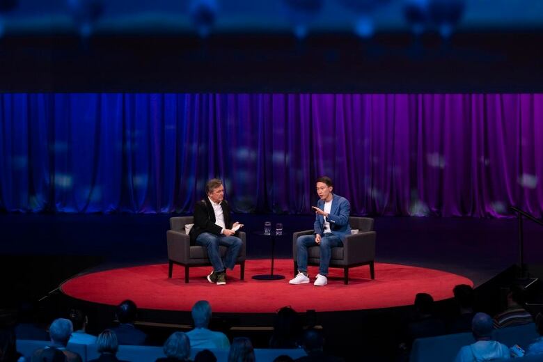 Two men in discussion sit in chairs sit on a stage.