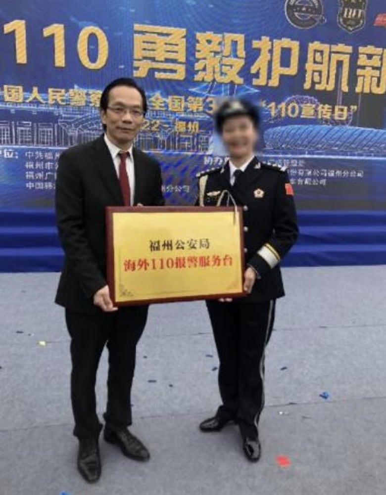 Man in suit holds plaque next to woman in uniform with blurred face.