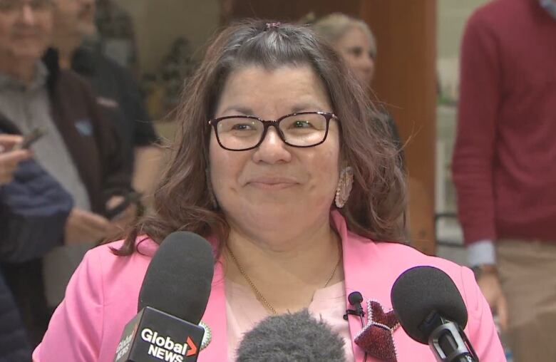 A woman with long brown hair and glasses wears a pink blazer with a pink shirt. She is standing at a podium with microphones.