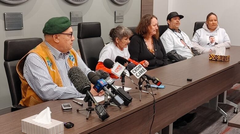 Several Indigenous people are sitting at a table in a conference room. They are all sitting in a line. The one in the far left is sitting in front of multiple microphones. A couple of tissue boxes sit on the table.