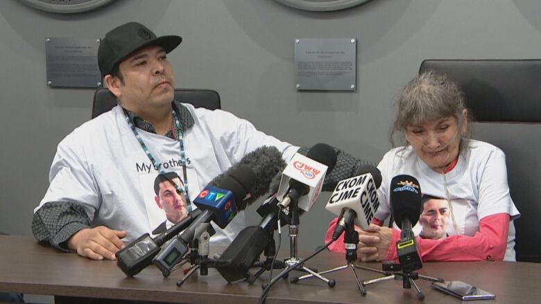 An Indigenous man wearing a black ball cap, lanyard, and white t-shirt over a dress shirt, is seated next to an elderly woman. His hand is soothing her back. The woman, who has grey hair, is wearing a matching white t-shirt over a pink longsleeve shirt. She has oxygen tubes running through her nose. She's crying while seated at a table, in front of multiple microphones.