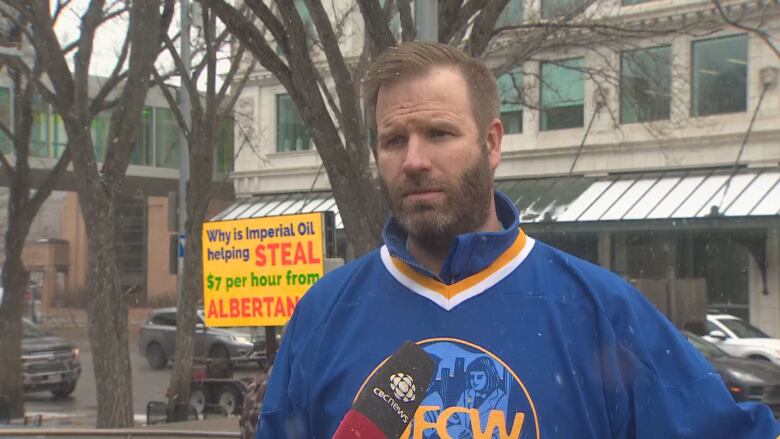 A man stands in front of a CBC News mic. In the background, a poster sign says 