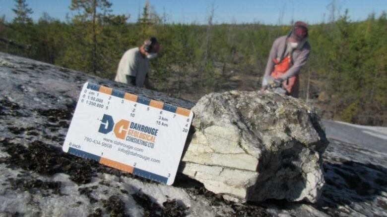 A rock sits on the ground with two men working in the background.