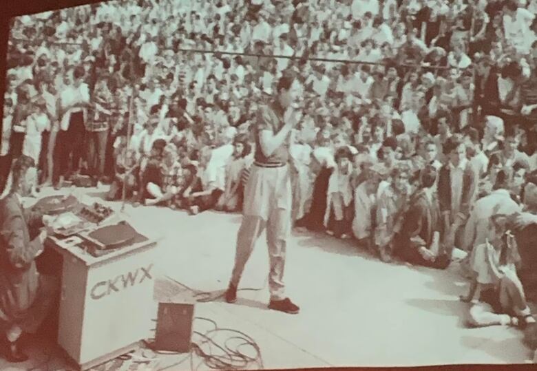 A black-and-white picture of a man holding a mic and talking to a group of assembled children.