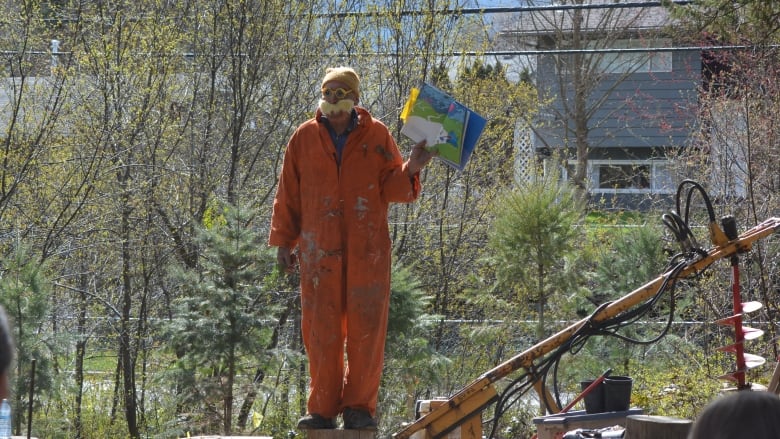 A man dressed in an orange jumpsuit with a large fake moustache and goggles reads a book to kids sitting outside under a large tree. 