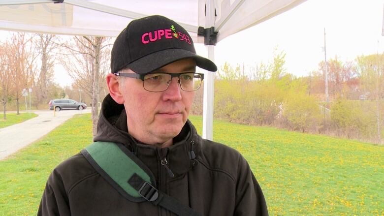 A man wearing a union hat, glasses, a black jacket and a green backpack standing under a white tent