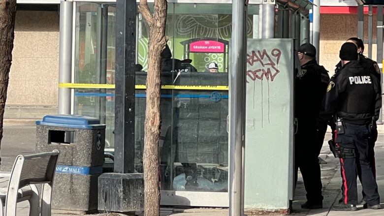 Three people dressed in black with the word POLICE across the back of their jackets, stand beside a bus shelter that has a yellow police tape wrapped around it.