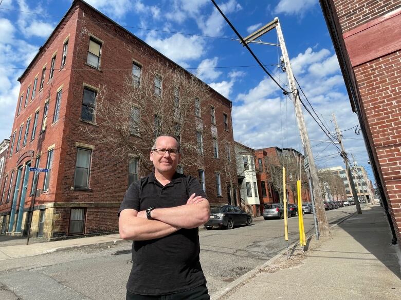 A man dressed all in black, with his arms crossed, standing on the sidewalk in front of a red brick building.