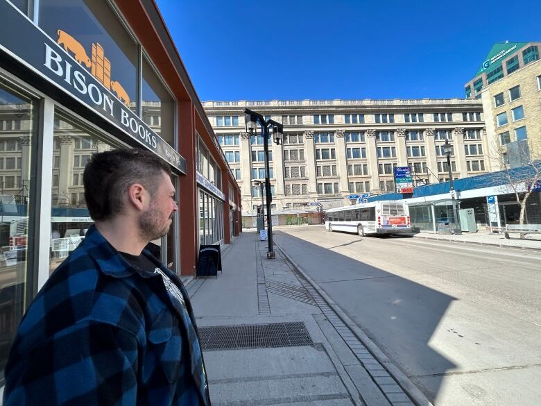 A person in a black and blue flannel standing in front of a book store stares across the street at a bus shelter where emergency crews found a body hours earlier.