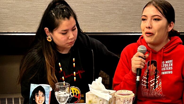 Woman in red hoodie speaks while holding a microphone while a woman in a black hoodie comforts her. 