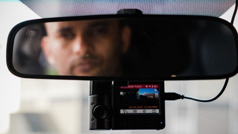 A close-up shows an Indian man in a rearview mirror, blurred, while an in-car video camera is seen with full focus.