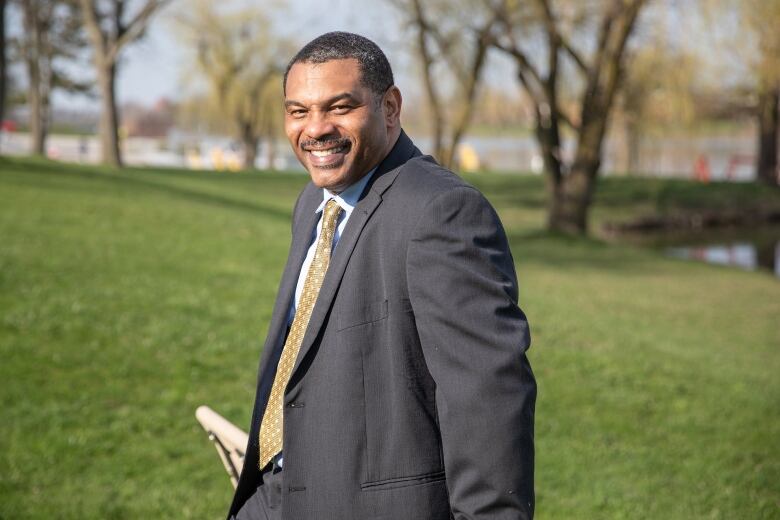 A Black man with short black hair and a mustache wearing a suit stands against a park bench and smiles at the camera.