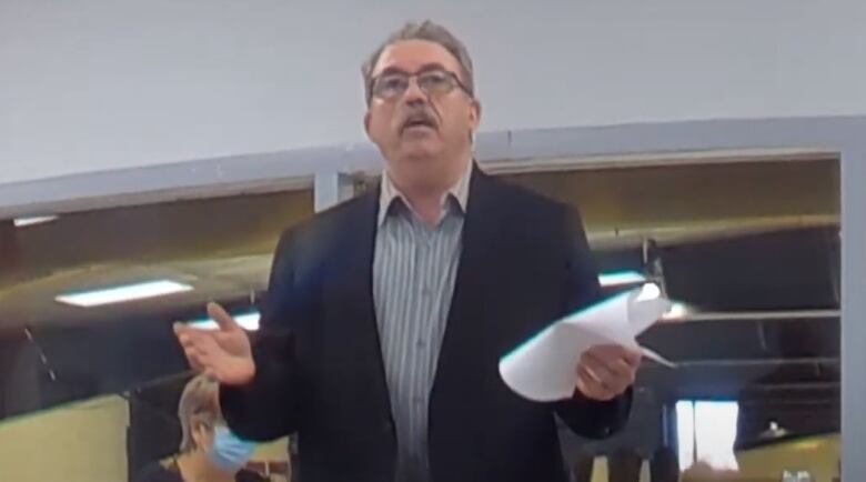 A man with grey hair and mustache and glasses wearing a blue blazer and striped shirt stands at a public meeting, holding his hands to his sides. 