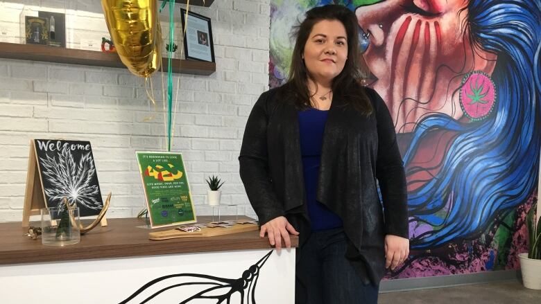 A woman stands beside a counter and looks toward the camera and smiles. Indigenous art is painted on the wall behind her.