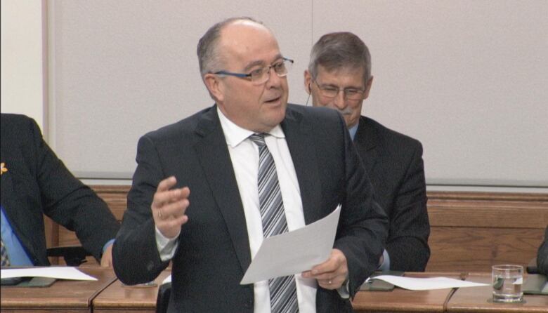 A man wearing a suit and holding a piece of paper speaks.