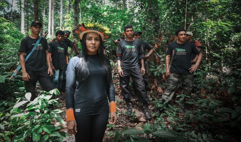 Ten people wearing all black stand in a forest, staring at the camera. The woman in the front wears a colourful headdress. 