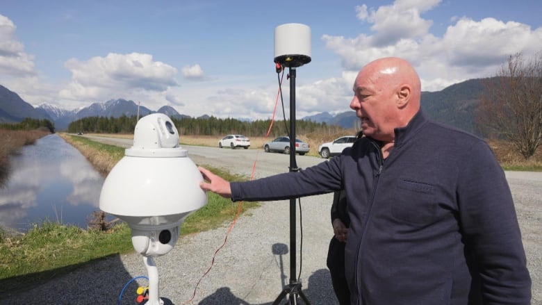 A man points to a white camera device used to detect drones. In the distance are a creek and mountains.