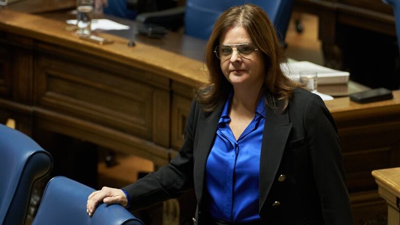 A woman in a black blazer and blue blouse stands in a legislative chamber.