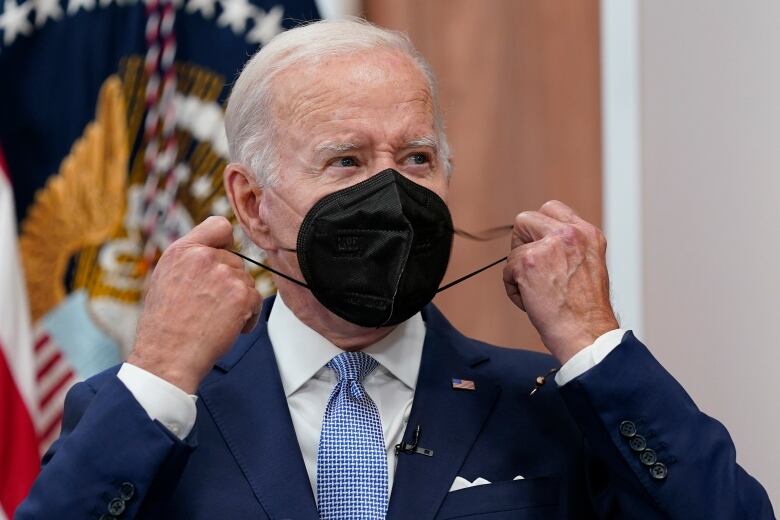 A white-haired man wearing a blue suit removes a black face mask.