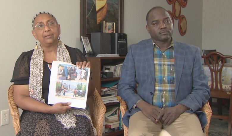 A woman and a man are seen seated beside each other. The woman is holding a piece of paper with images of the damaged buildings in Khartoum.